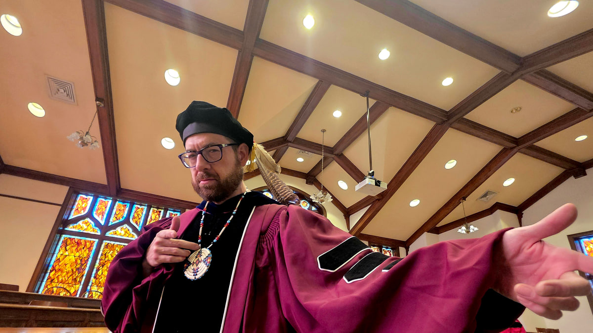Darren Lone Fight in academic regalia and a beaded medallion, gesturing in a wood-beamed room with stained-glass windows.