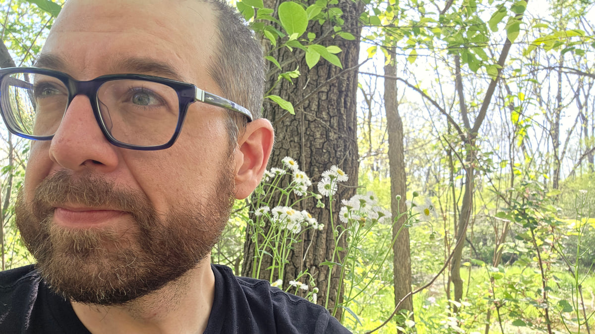Darren Lone Fight in glasses beside a tree and wildflowers in a sunlit wooded setting.